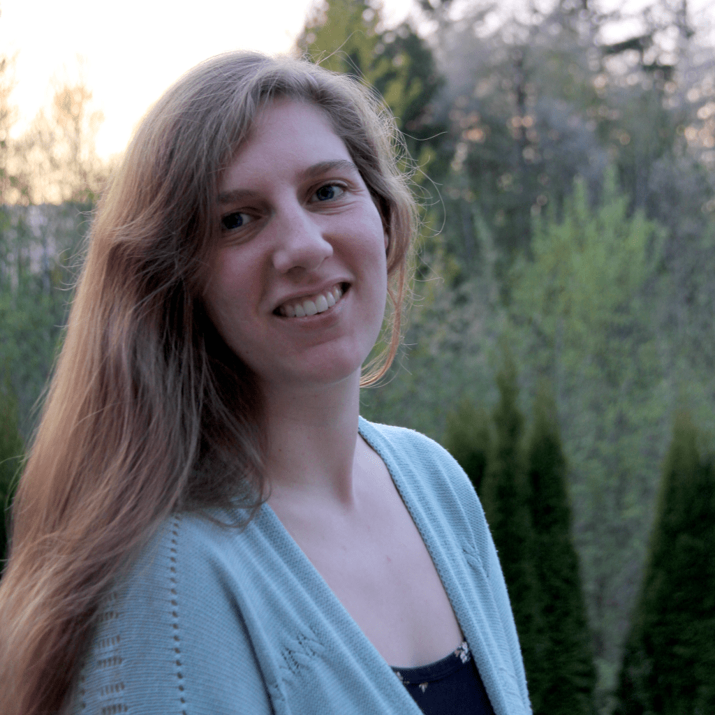 Headshot of a woman with long hair, wearing a blue cardigan with trees in the background.